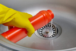 A gloved hand pouring a liquid drain cleaner down a sink drain.