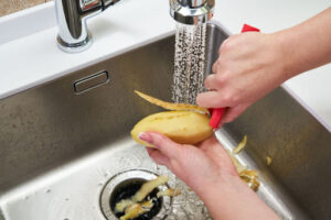 Someone peeling a potato in a kitchen sink with a garbage disposal and running water.