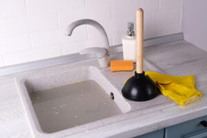 A clogged sink with standing water. There's a plunger, gloves, sponge, and soap bottle on the countertop.