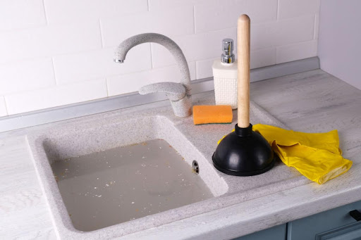 A clogged sink with standing water. There's a plunger, gloves, sponge, and soap bottle on the countertop.