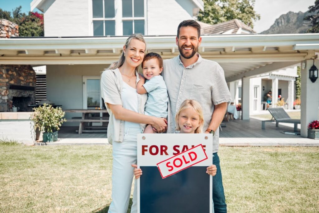 A family of four standing in front of the house they bought with a "for sale" sign that now says "sold."